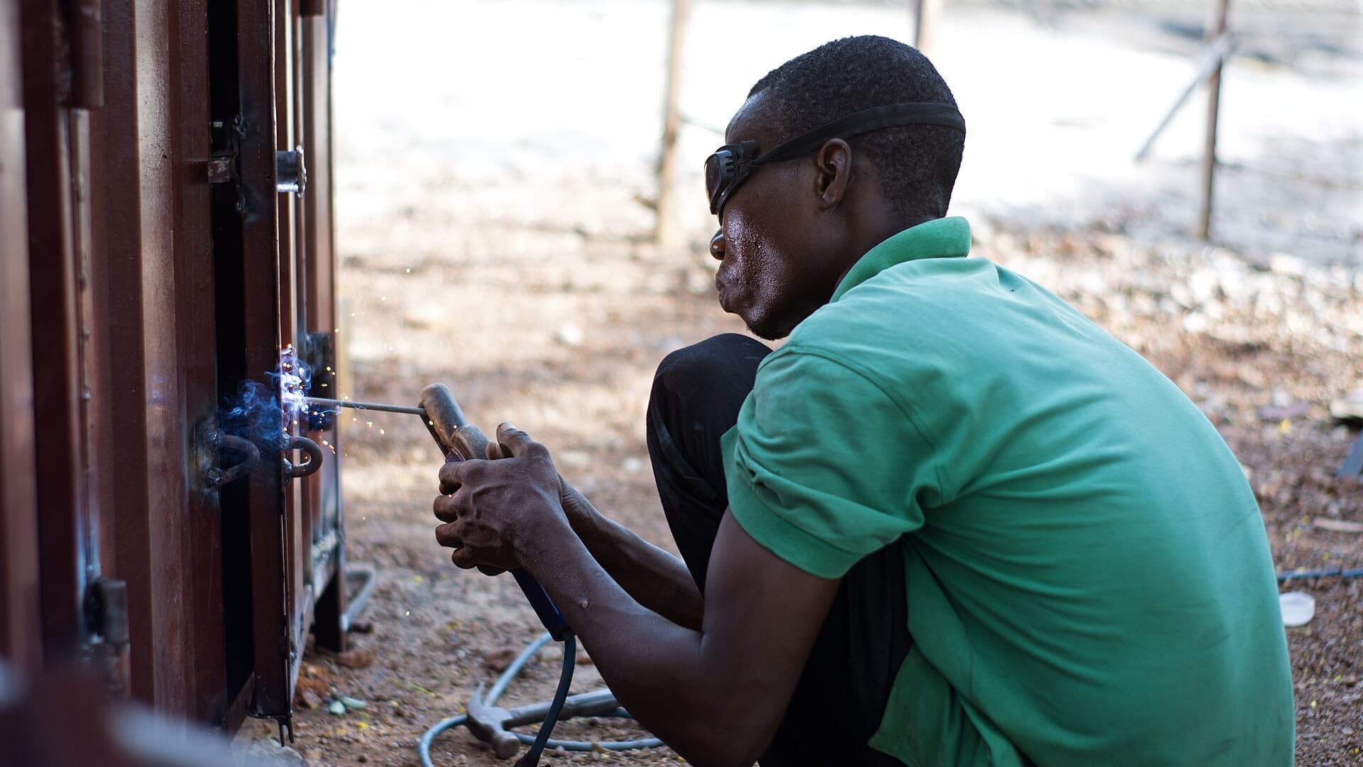 A welder in a green shirt joining a metal gate frame, sparks flying, working outdoors on red earth.